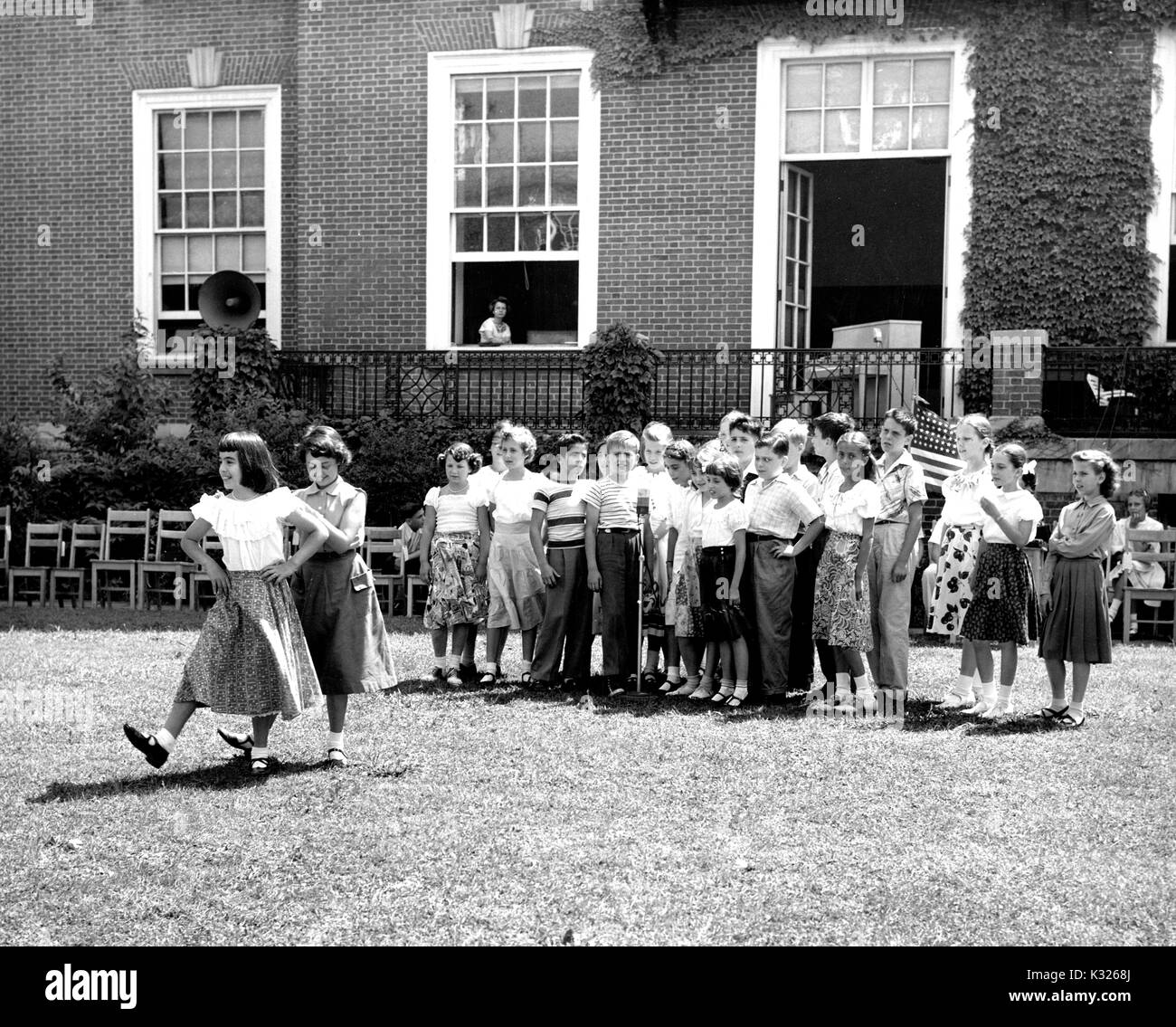 At the end of the school year for a demonstration school at Johns Hopkins University, boys and girls put on a show in the grass on a sunny day, standing in a line behind a microphone while two girls wearing skirts show off their dance moves, outside of an ivy-covered campus building, Baltimore, Maryland, July, 1950. Stock Photo