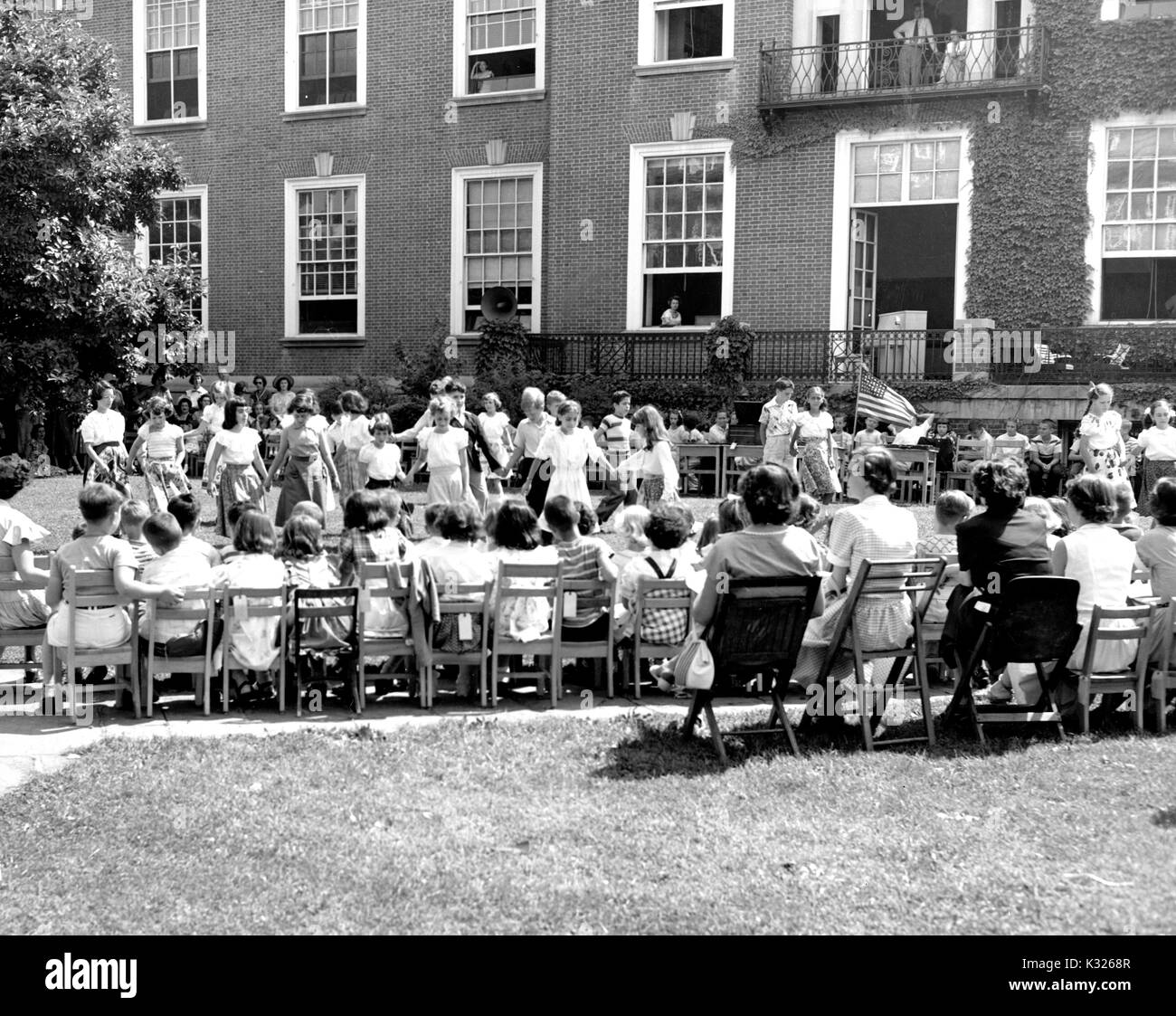 At the end of the school year for a demonstration school at Johns Hopkins University, young boys and girls put on a show in the grass on a sunny day, standing in a group holding hands while one girl waves an American flag, in front of an audience made up of parents, teachers, and classmates sitting in chairs outside of an ivy-covered campus building, Baltimore, Maryland, July, 1950. Stock Photo