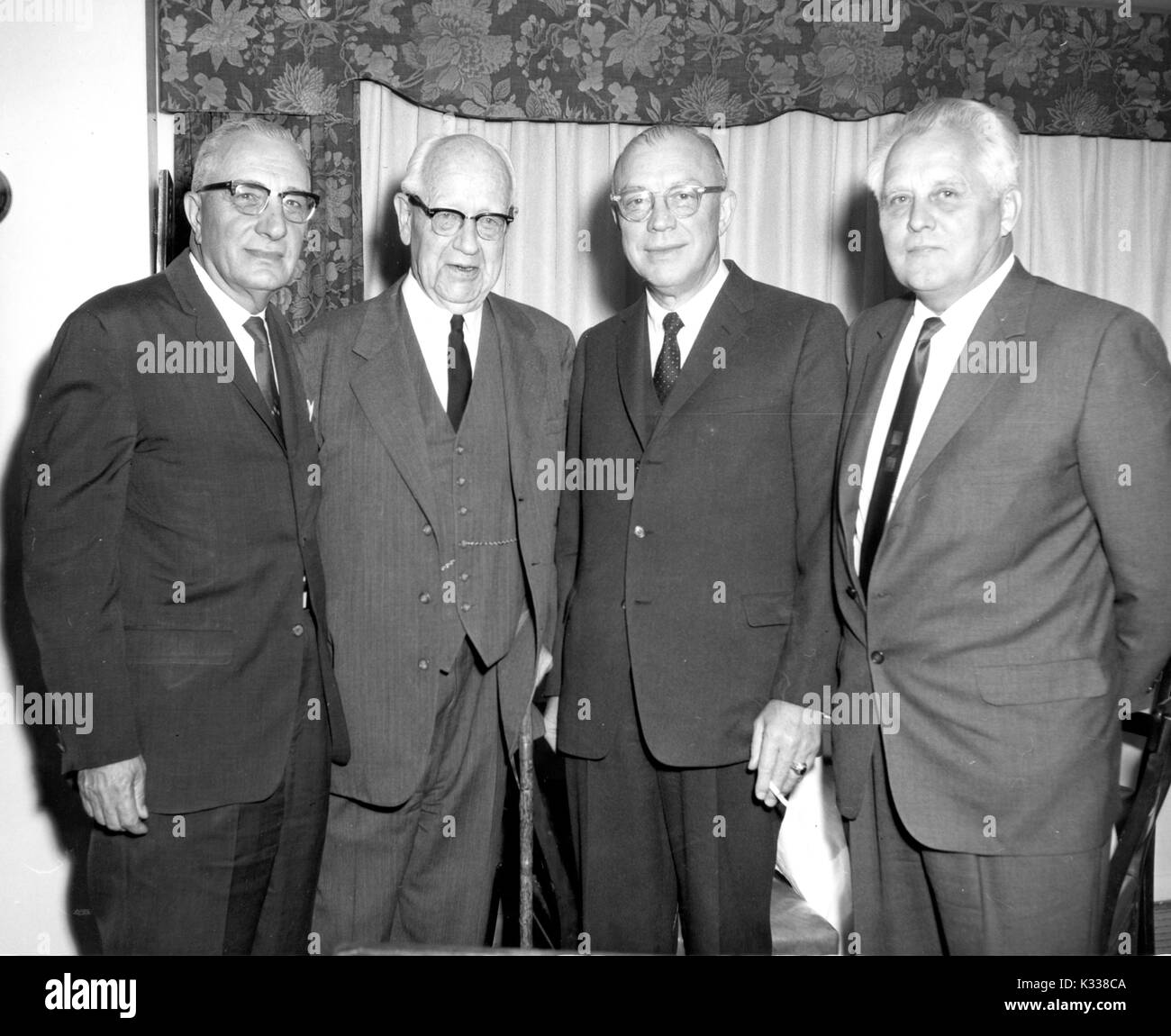 Milton Stover Eisenhower -- President of Johns Hopkins University -- smokes a cigarette while standing to pose for a photograph with Congressmen, everyone wearing suits and all but one in glasses, including Congressmen Harold Ostertag from New York and James Auchincloss from New Jersey, next to famous portrait artist Bjorn Egeli, Baltimore, Maryland, 1966. Stock Photo