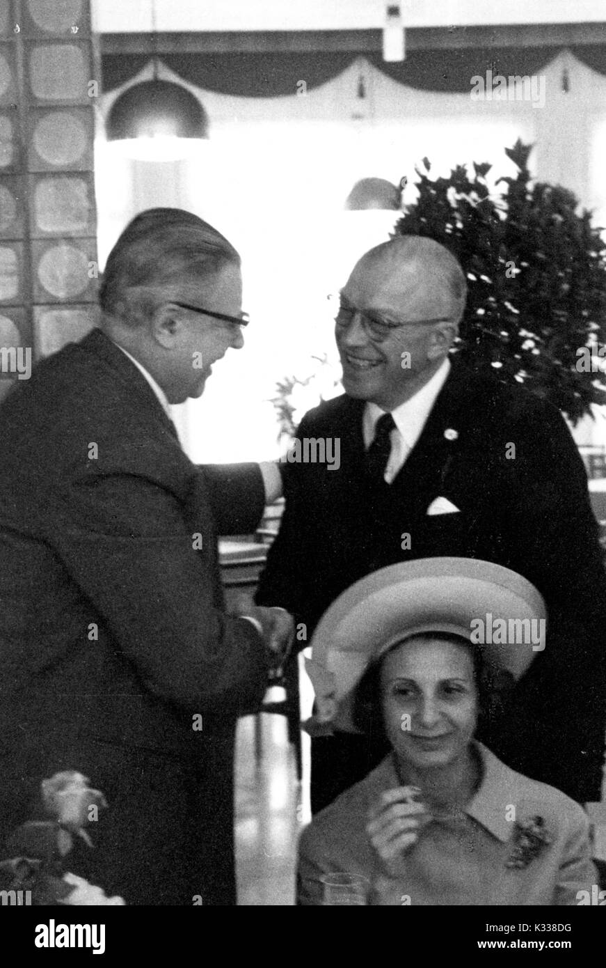 During the Associazione Italo-Americana Luncheon at the School of Advanced International Studies (SAIS) in Washington, DC, President of Johns Hopkins University Milton Stover Eisenhower -- around 62 years of age -- smiles and stands to shake hands with another guest in attendance, while Eisenhower's wife sits in front of them wearing a hat and smoking a cigarette, smiling, Washington, DC, 1965. Stock Photo