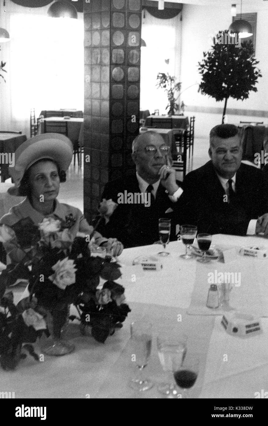 During the Associazione Italo-Americana Luncheon at the School of Advanced International Studies (SAIS) in Washington, DC, President of Johns Hopkins University Milton Stover Eisenhower -- around 62 years of age -- sits at a table holding his hand to his chin, with his wife to his right wearing a hat and smoking a cigarette, and another guest to his left and wine glasses spread around a bouquet on the table, Washington, DC, 1965. Stock Photo