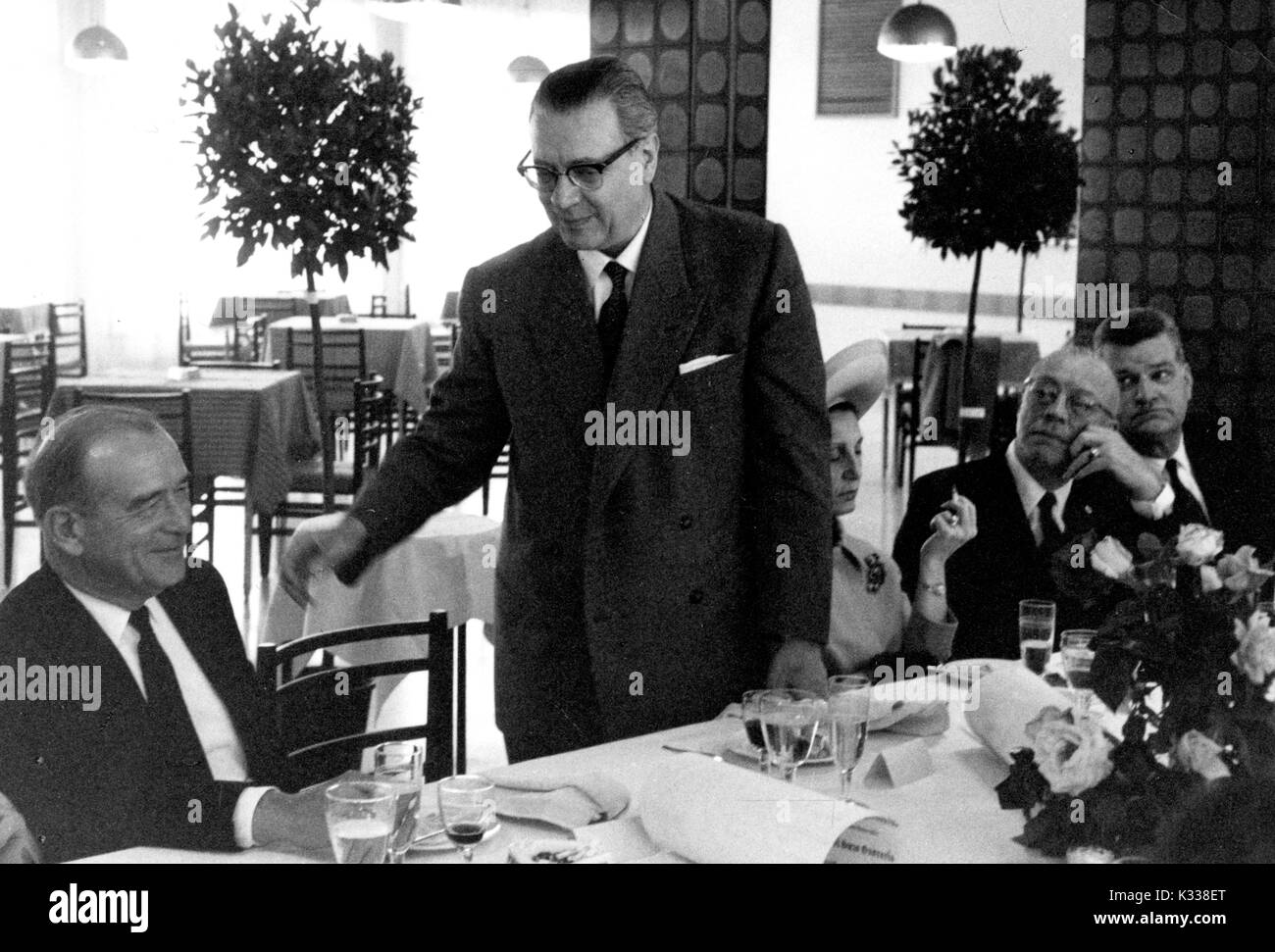 During the Associazione Italo-Americana Luncheon at the School of Advanced International Studies (SAIS) in Washington, DC, President of Johns Hopkins University Milton Stover Eisenhower -- around 62 years of age -- sits at a table with his wife to his right, while other luncheon guests take their seats, one gentleman standing in the middle, other tables and trees in the background, and plates and flowers on the table in front, Washington, DC, 1965. Stock Photo