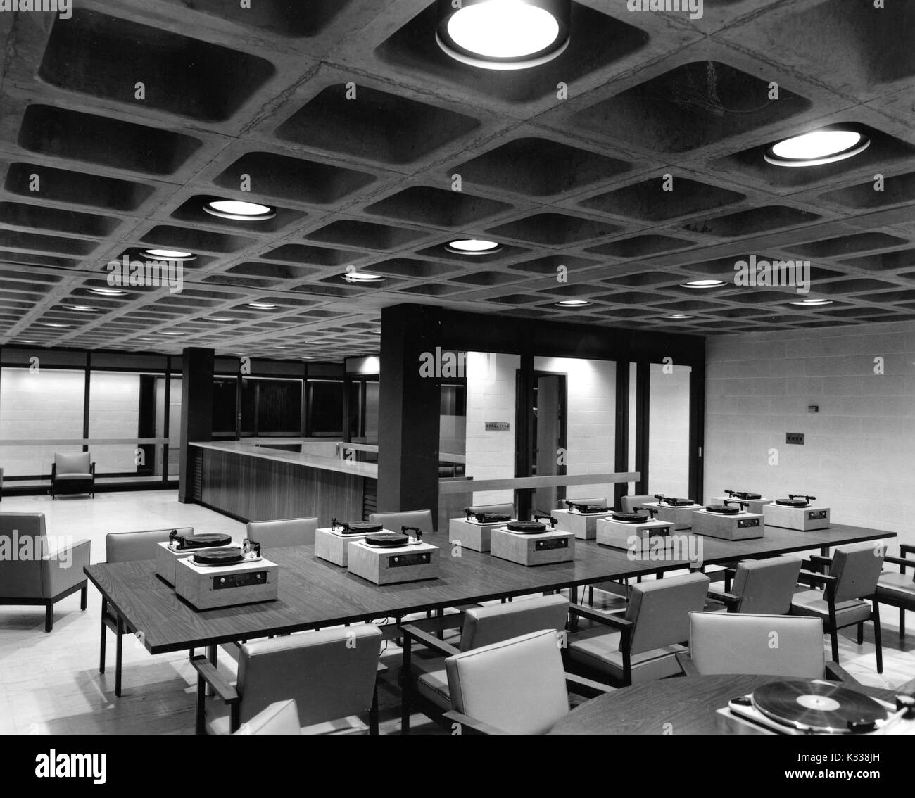 Inside the Milton S Eisenhower Library at Johns Hopkins University, the audiovisual center includes a long table with multiple record players and empty chairs, in front of a counter in the back for checking out materials, and bright lights on the ceiling, Baltimore, Maryland, 1965. Stock Photo