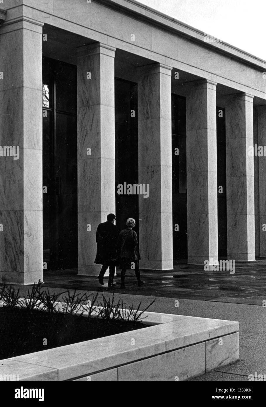 The entrance of newly opened Milton S Eisenhower Library at Johns Hopkins University from the terrace level, with a man and a woman walking side by side into the glass doors on Q Level in between the tall marble columns, with a small plot of shrubs in the foreground, on a rainy day, Baltimore, Maryland, December 12, 1964. Stock Photo