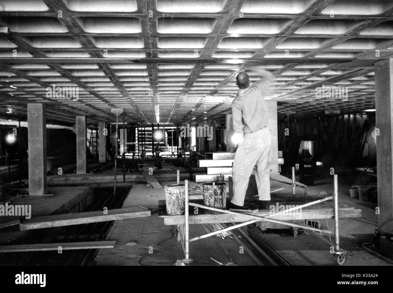 During the early stages of construction of the Milton S Eisenhower Library at Johns Hopkins University, a construction worker wearing a uniform stands on a platformn with his back turned inside an unfinished M Level, painting glaze onto the beams of the ceiling, with beams and wooden boards scattered around the inside of the building, and a courtyard visible in the distance with sunlight pouring in from the terrace level, Baltimore, Maryland, November 13, 1963. Stock Photo