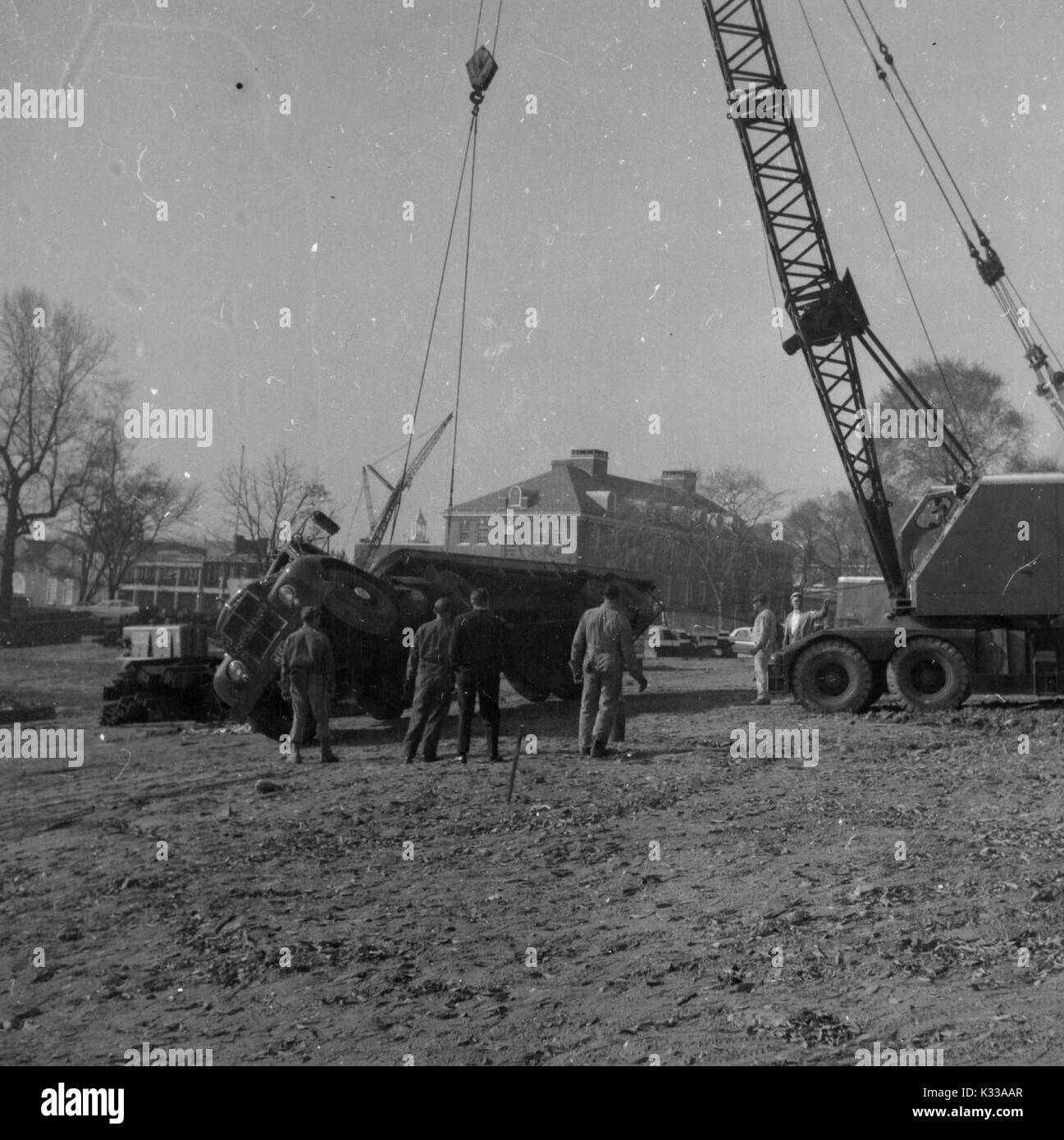 During the early stages of construction of the Milton S Eisenhower Library at Johns Hopkins University, a half dozen workers in uniform use a large crane to attempt to correct an overturned truck to its upright position, on the muddy grounds in front of Remsen Hall, a chemistry building on campus, Baltimore, Maryland, 1963. Stock Photo