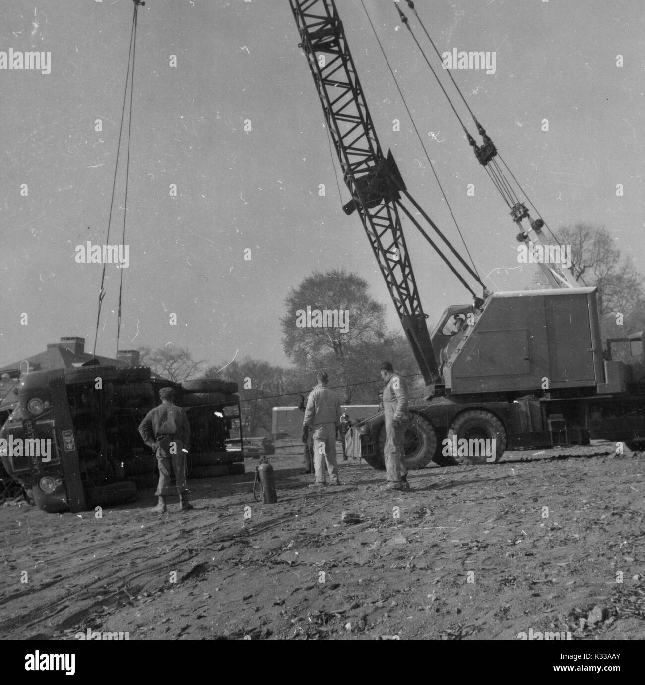 During the early stages of construction of the Milton S Eisenhower Library at Johns Hopkins University, an overturned drunk is lifted back to its upright position by a large crane, while three workers in boots and uniforms stand to oversee the process, above a rocky and muddy ground, Baltimore, Maryland, 1963. Stock Photo