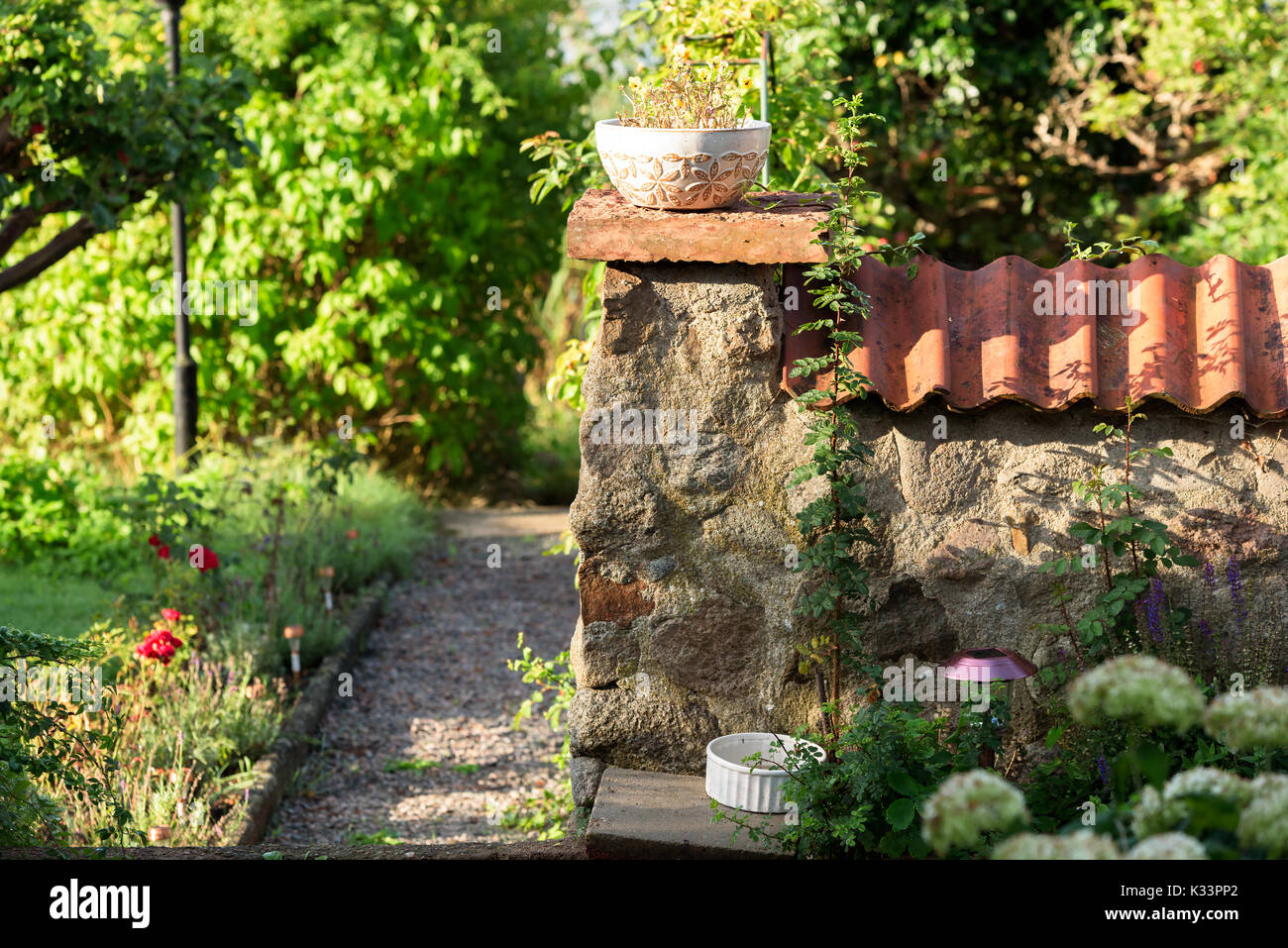 Corner of stone wall with flowerpot on top and a gravel garden path beside. Wall has roof tiles on top. Stock Photo
