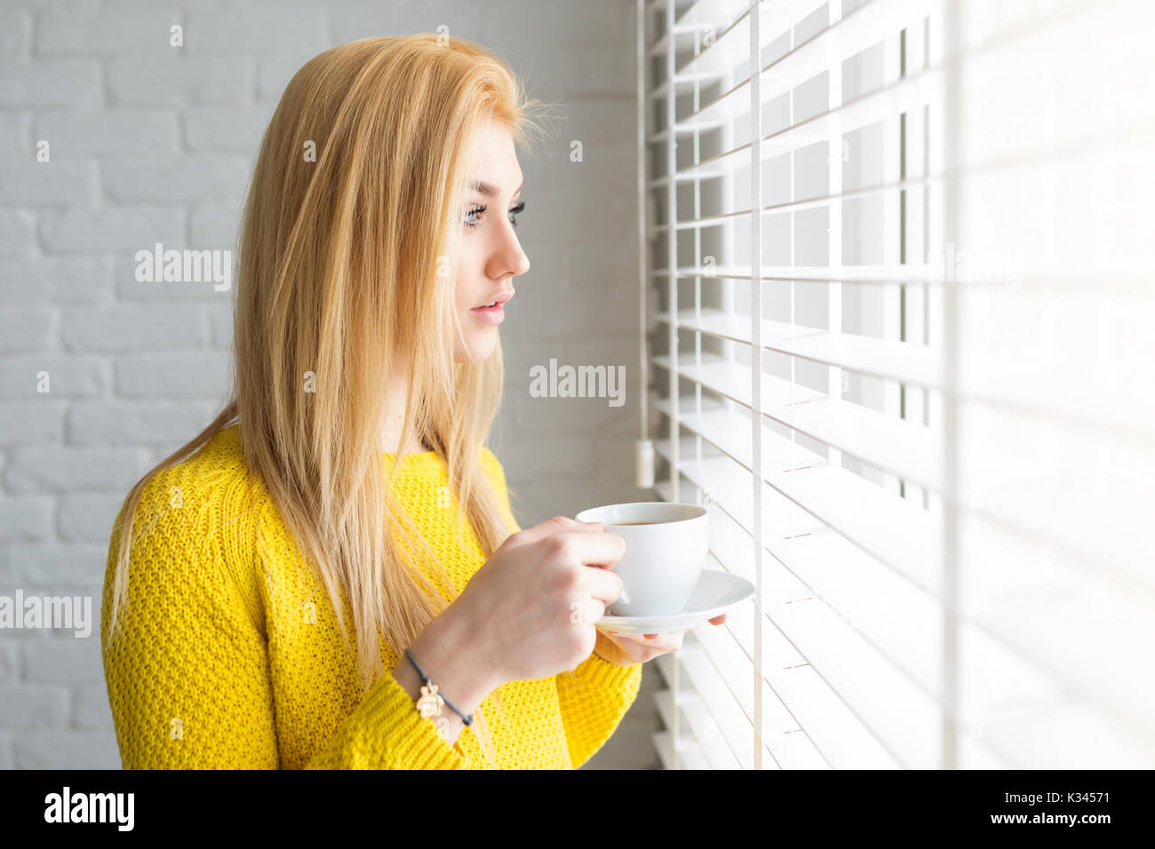 A photo of young woman looking outside the window. She's holding a cup of tea. Stock Photo