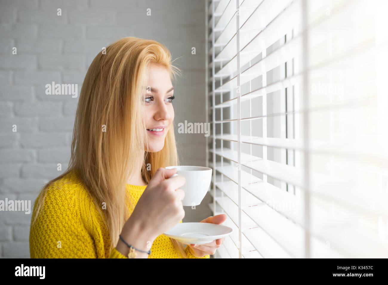A photo of young, beautiful woman with a cup of tea. She's looking outside the window and smiling. Stock Photo