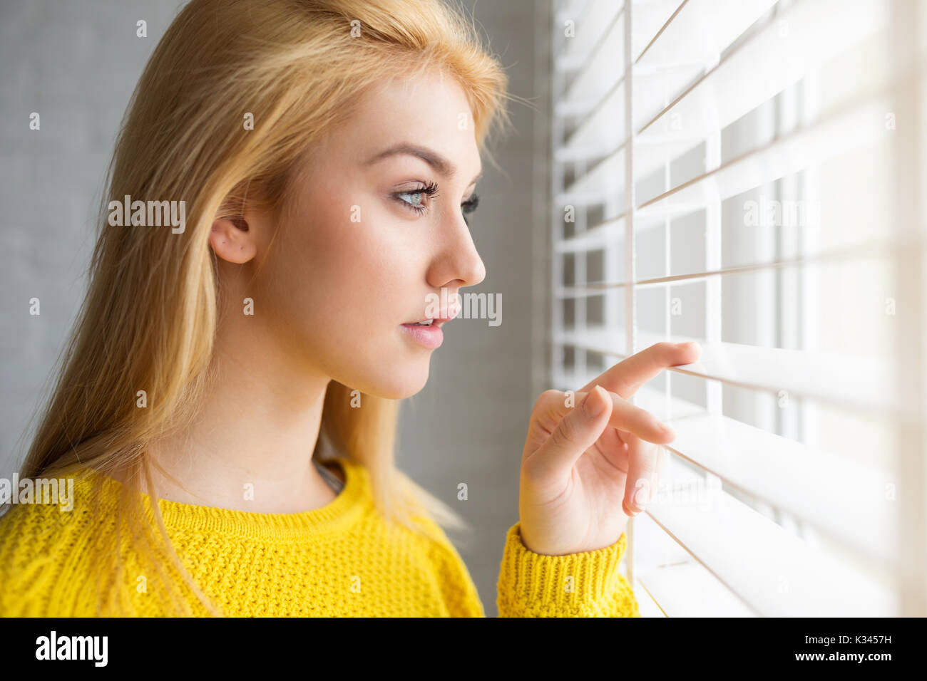 A photo of young, beautiful woman standing by the window and looking outside. She's waiting. Stock Photo