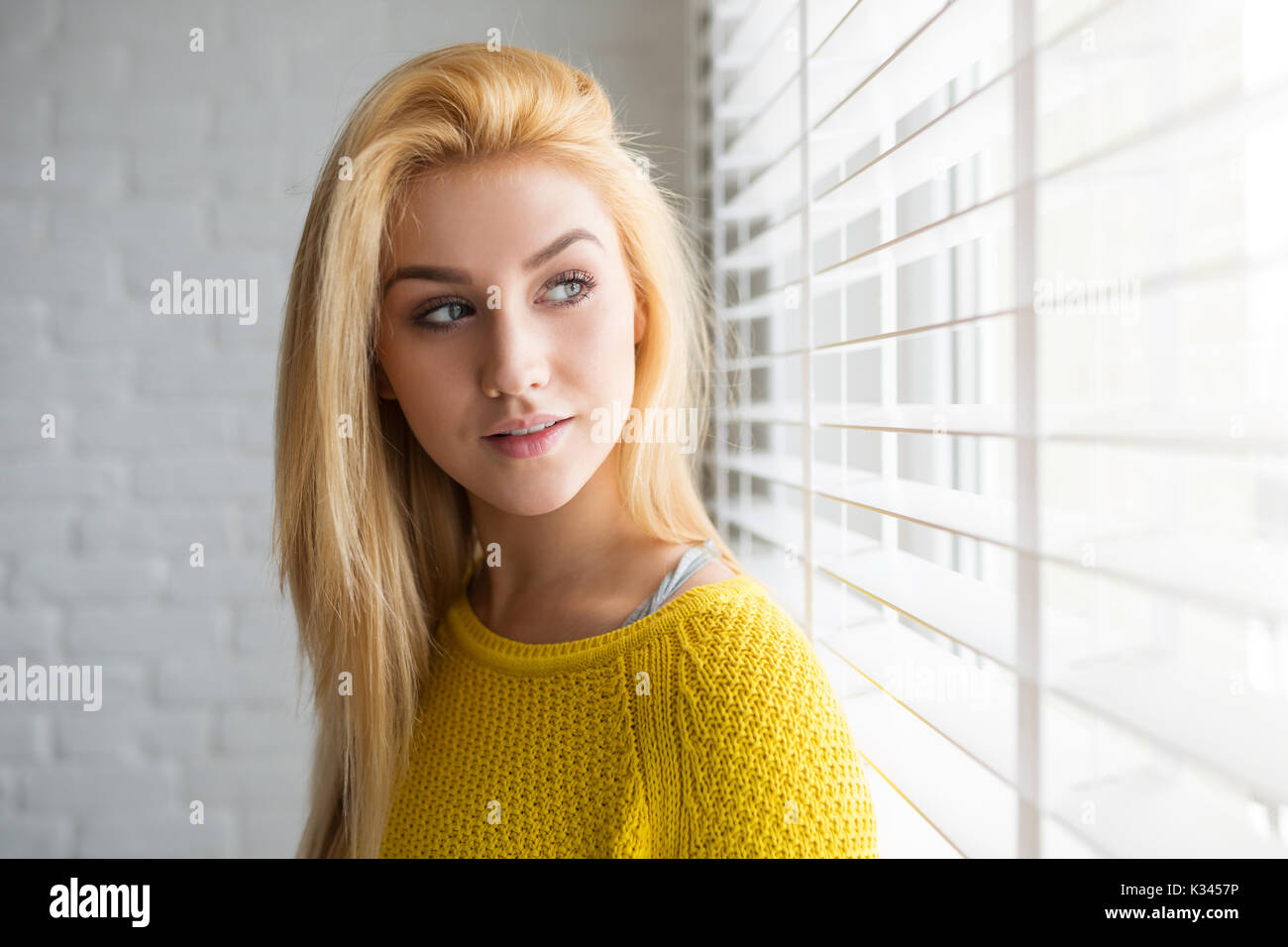 A photo of young, beautiful woman standing by the window. She's looking outside and waiting for something. Stock Photo