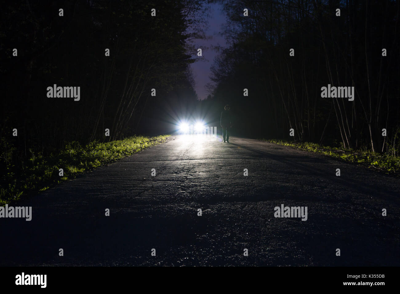 Male silhouette at the edge of a dark mountain road through the forest in the night. Scenic night landscape of dark blue sky. Man standing on the road Stock Photo