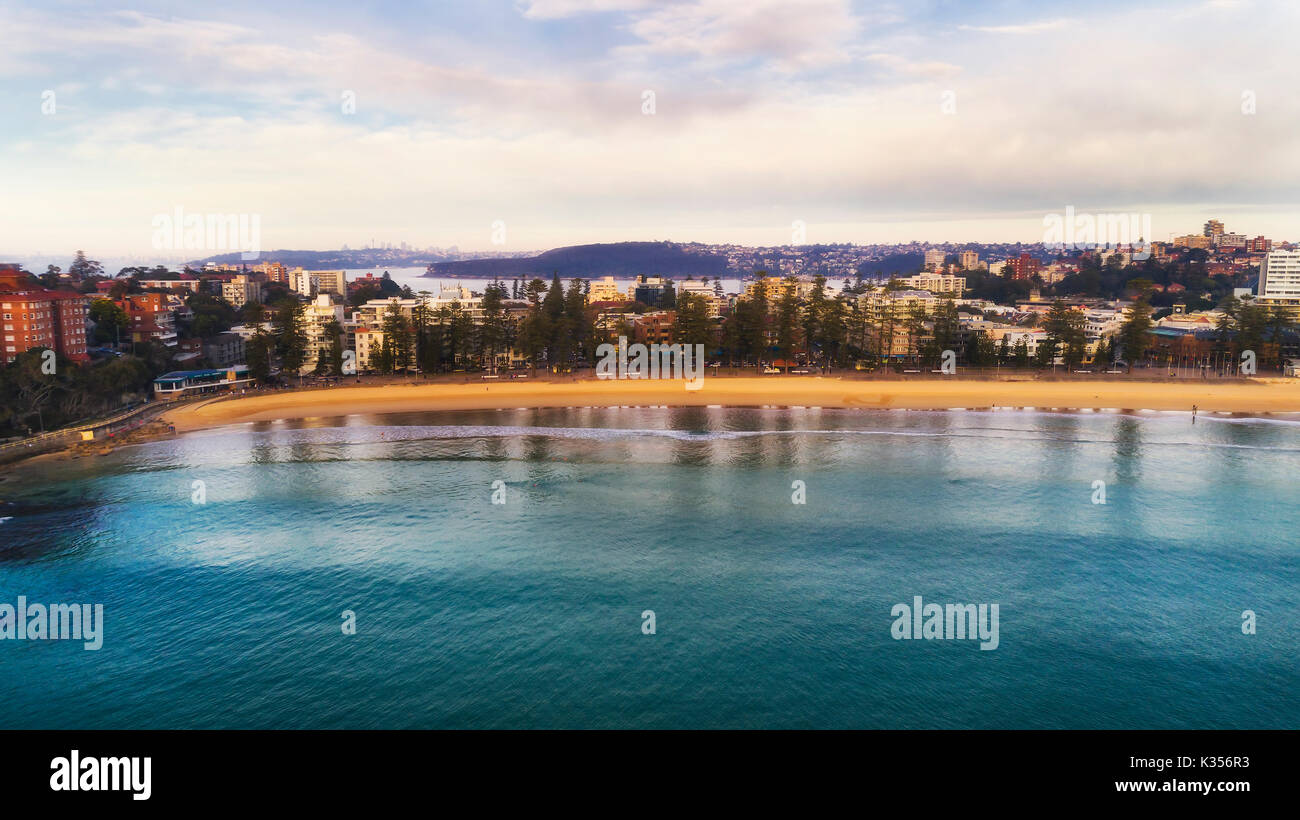 Frontal waterfront of sandy Manly beach from mid-air drone flight towards inland middle harbour and distant CBD and middle head. Stock Photo