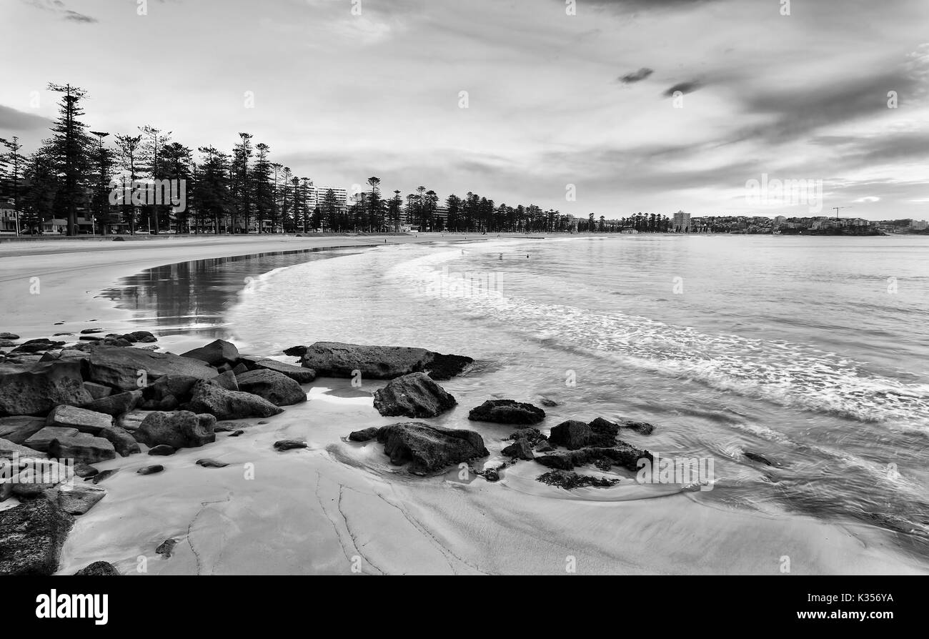 Flat waterfront of sandy Manly beach on Sydney Northern beaches with smooth blurred wave slowly rolling to coastal boulders during low tide. Stock Photo