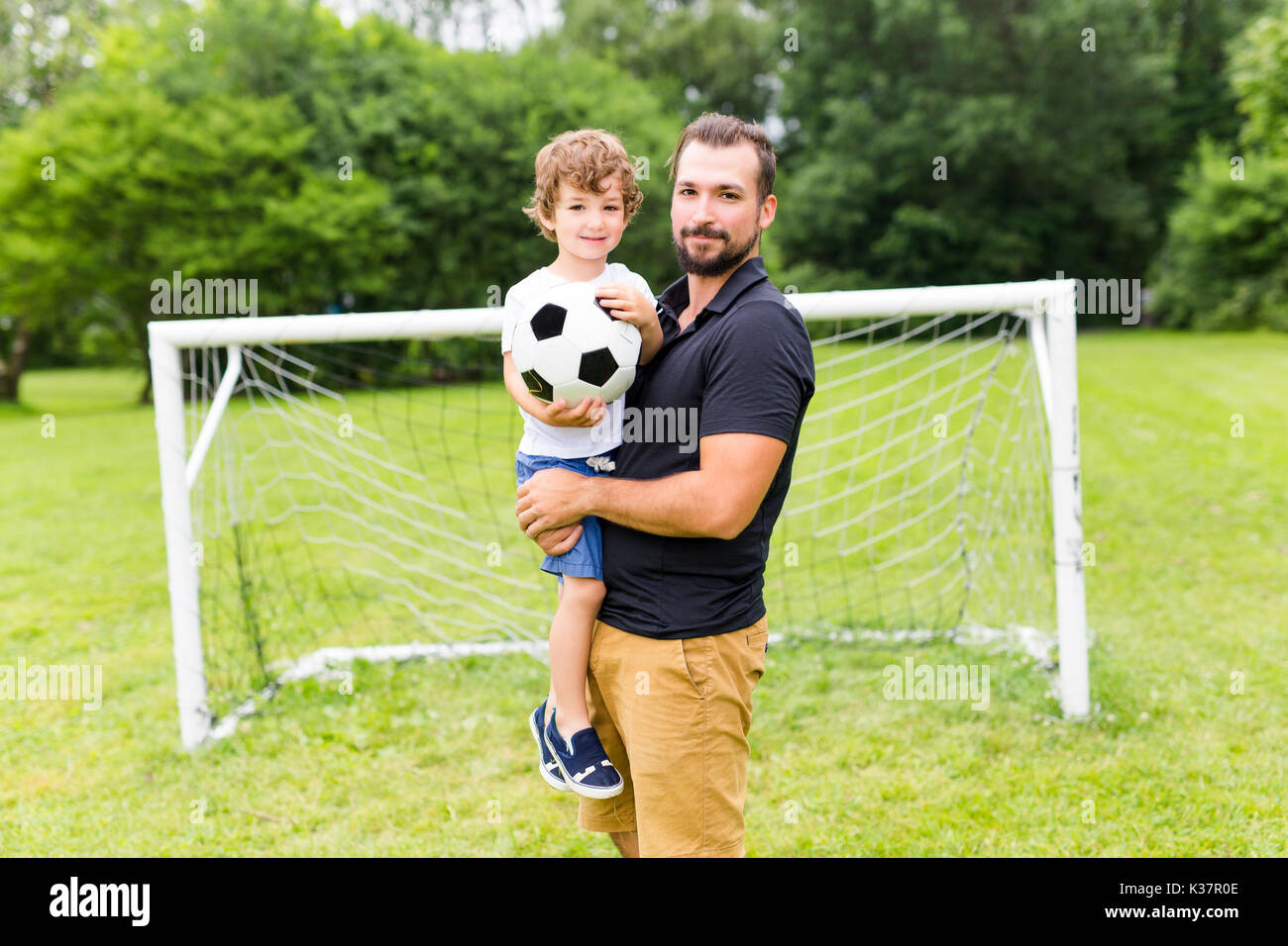 father with son playing football on football pitch Stock Photo