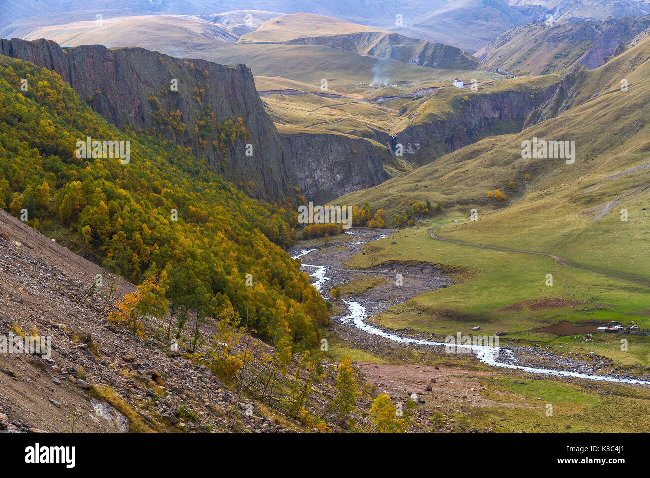 Autumn mountain landscape: hill slopes covered by dry plants and trees with yellow and green foliage, valley with a river and mountains in fog at the  Stock Photo