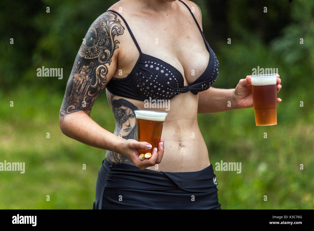 A young tattooed woman carries two beers in plastic cups, summer scenery, Czech Republic young thirties Stock Photo