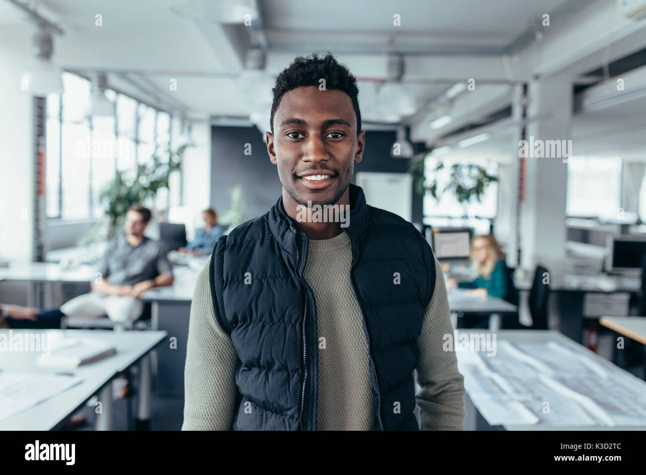 Portrait of happy young businessman standing in office with people sitting in background. African male designer standing in modern office. Stock Photo