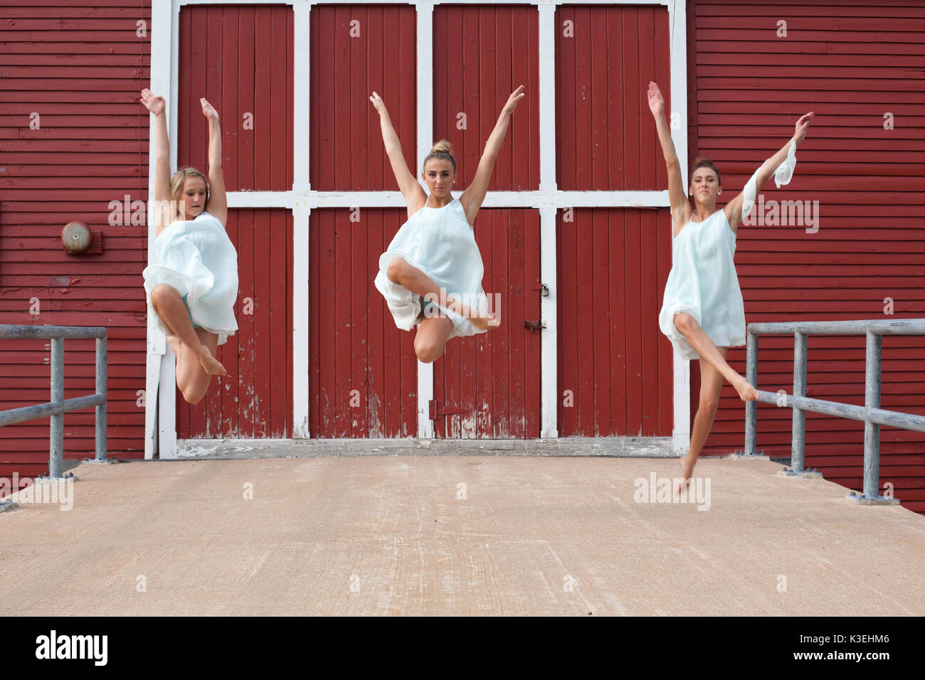 Three sisters in matching white dresses dancing outside red barn on