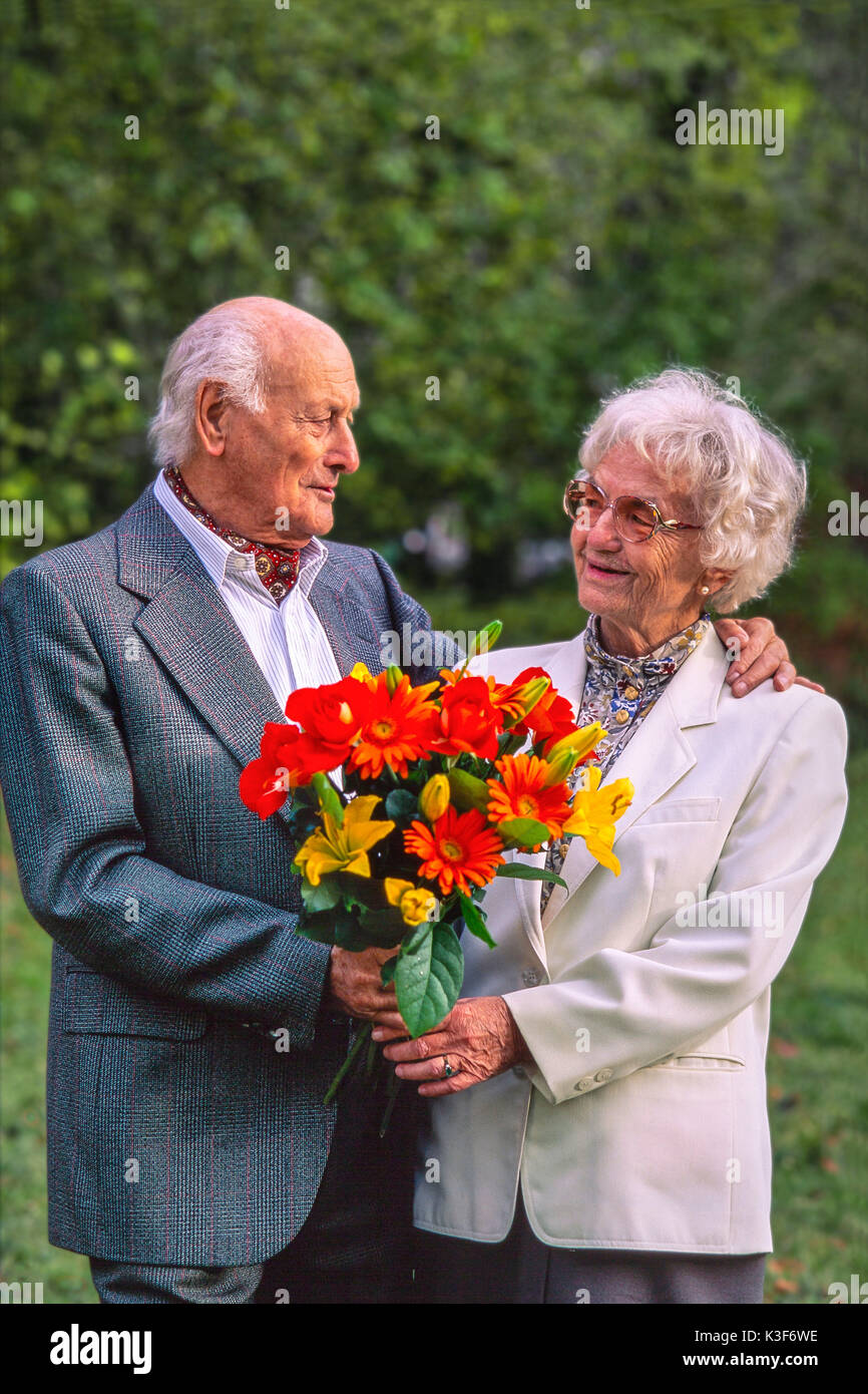 Senior citizen's couple with bouquet, the man has placed his arm around the woman and looks at them Stock Photo
