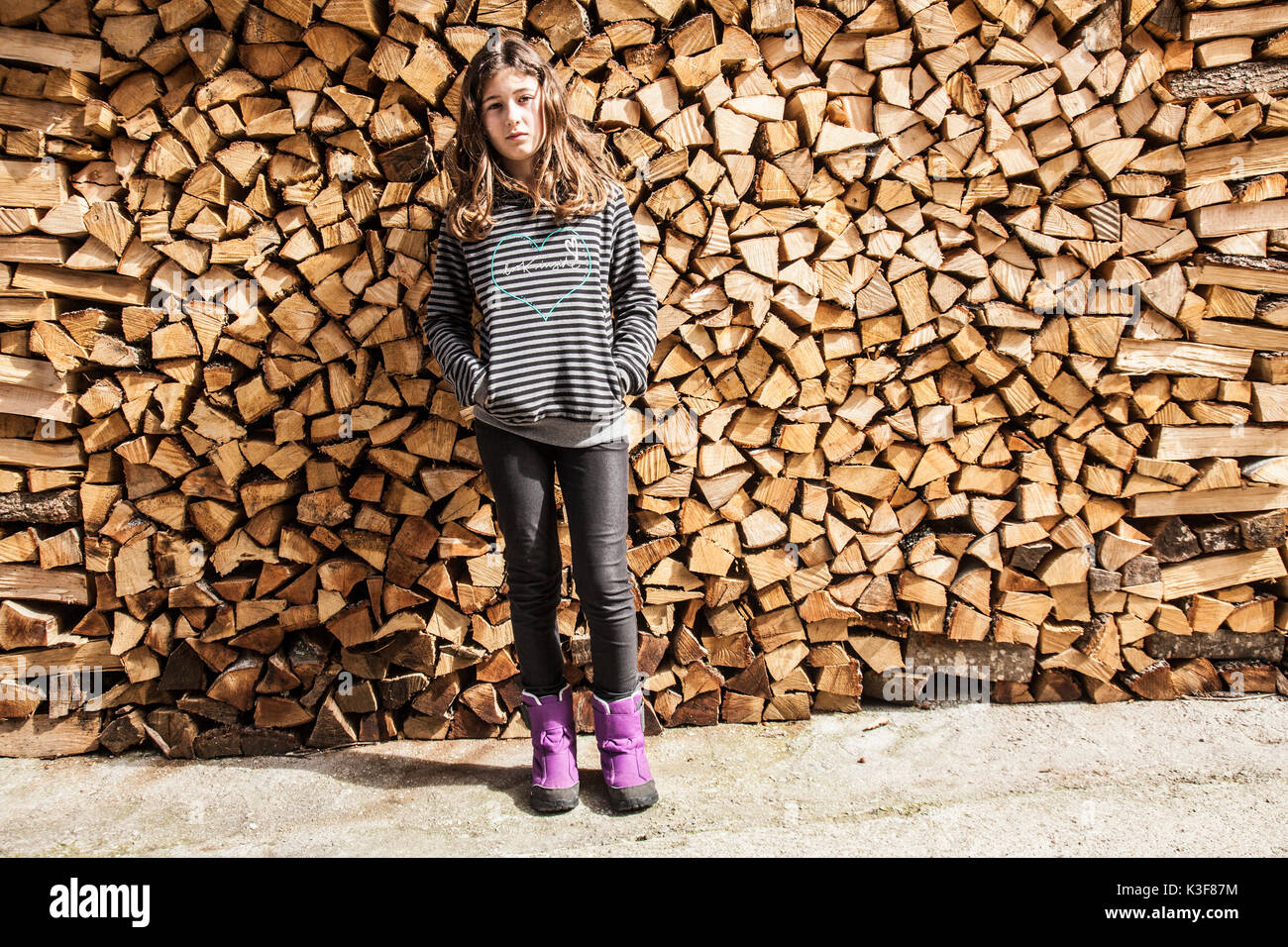 Full-Length Portrait of Serious Girl Standing in Front of Firewood Stock Photo