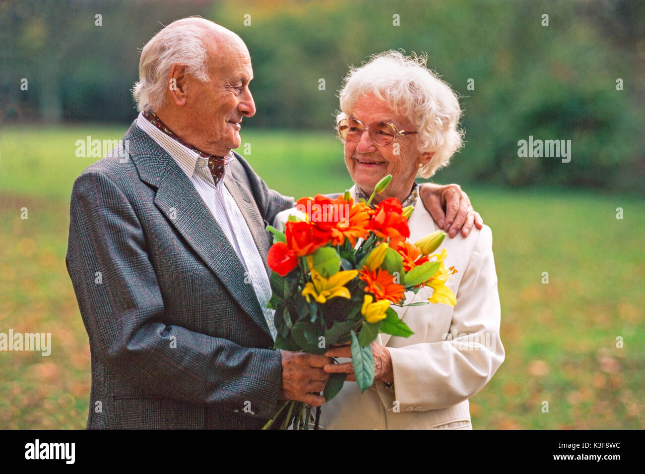 Senior citizen's couple with bouquet, the man has placed his arm around the woman and looks at her Stock Photo