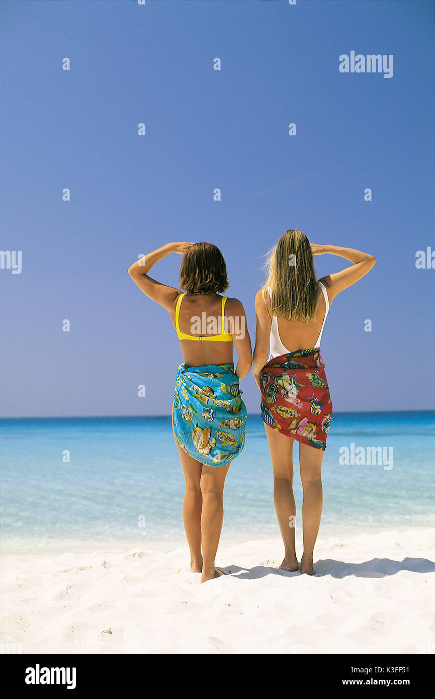 Two women to be seen from the back stand at the beach and see at the direction of horizon Stock Photo