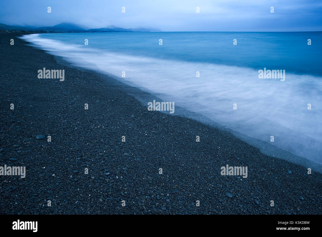 Long time exposure on the beach of Kaikoura, New Zealand. The black beach, blue sea, rain mood and in the background mountains prove a dark light mood. Stock Photo