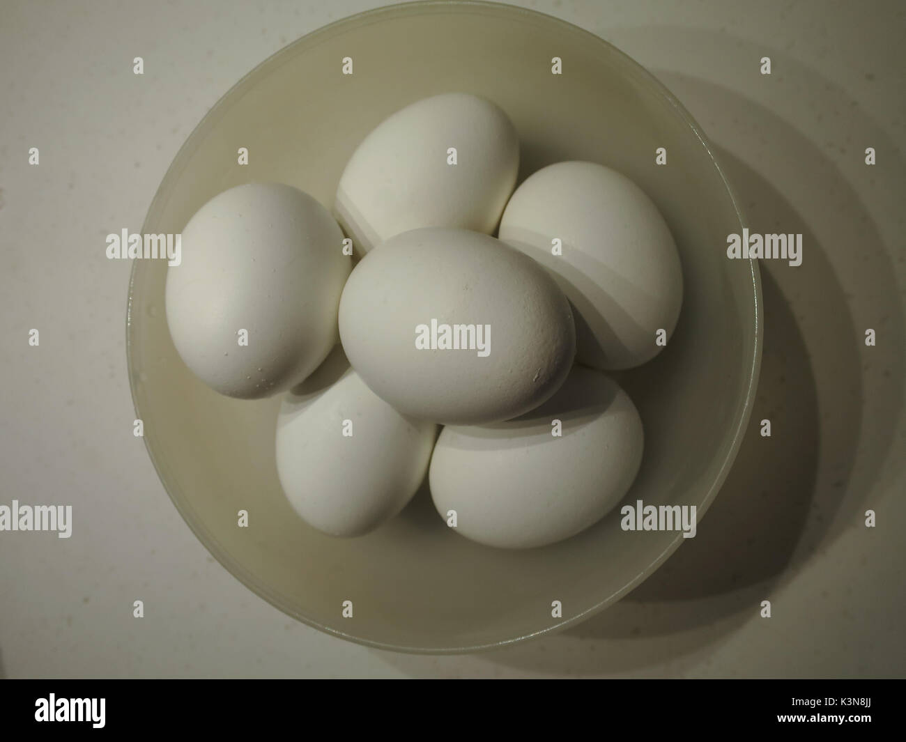 Bowl with six white chicken eggs, casts an interesting triplicate shadow, the eggs very white against a cream-colored bowl, full frame and unusual . Stock Photo