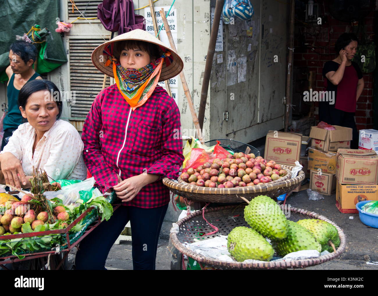 HANOI, VIETNAM - MAY 22, 2017: Woman selling tropical fruit from bicycle stalls on a busy Hanoi street fruit market wearing Vietnamese conical hats. Stock Photo