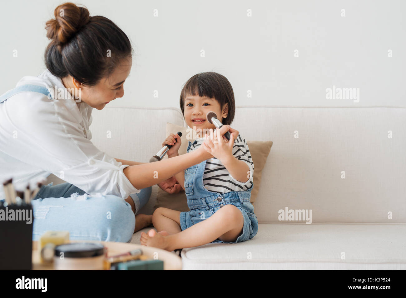 Cute little girl and her beautiful mother are doing makeup while sitting on couch at home Stock Photo