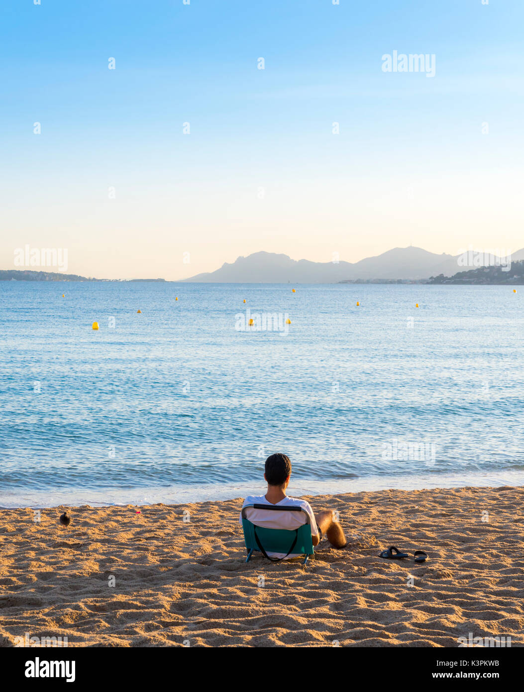 A young adult sits on an empty beach lounger overlooking a bay in Cote d'Azur, France at sunset Stock Photo
