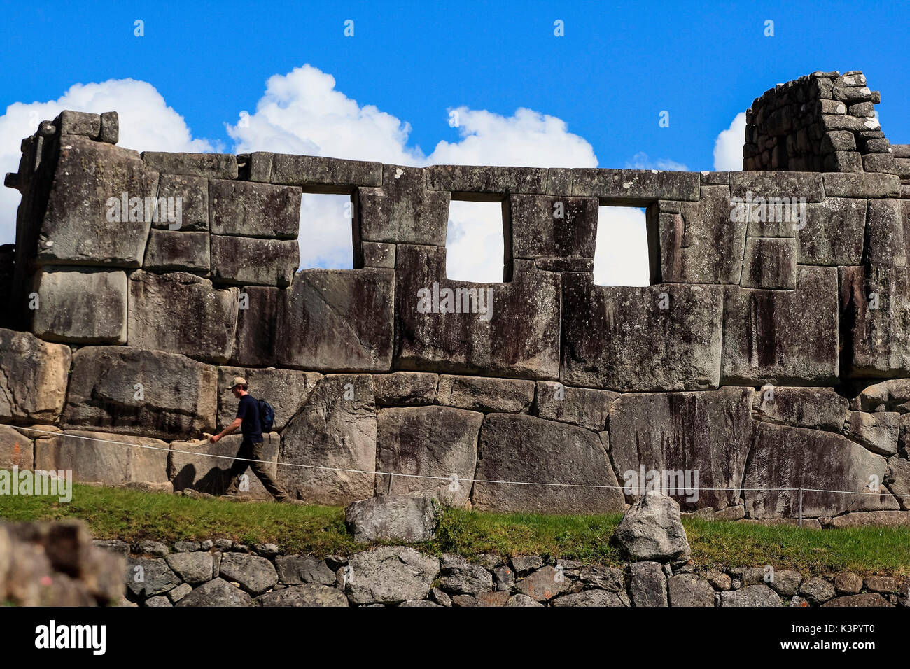 A tourist walking around the Inca ruins of Machu Picchu by the Temple of the Three Windows, consisting of only three walls on a rectangular base and covered by a roof made of adobe walls constructed from large blocks of solid rock carved in polygonal shape forming a conglomerate of perfectly matched stones each other, leaving space for, originally five windows, although there are today only three of them indicating the exact location of the sunrise Peru South America Stock Photo