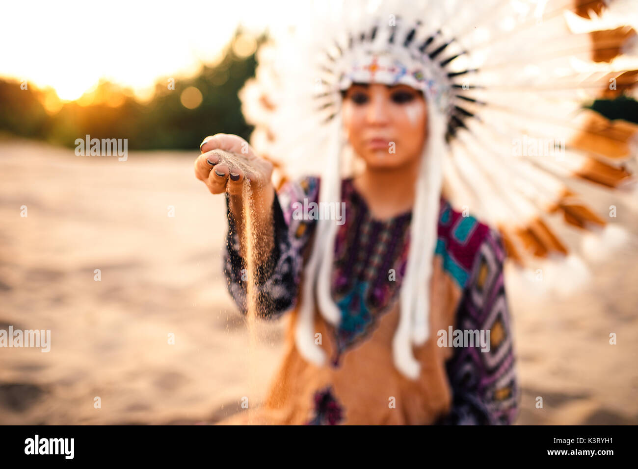 Beautiful woman in a suit of the American Indian play with sand on sunset. Young girl in headdress made of feathers of wild birds Stock Photo