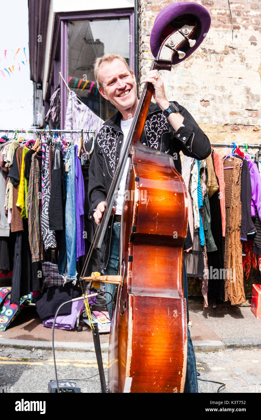 Celloist playing cello in the street during a concert by the skiffle band, High Chaparrals. Eye-contact, smiling. Daytime. Hat hanging on Cello. Stock Photo