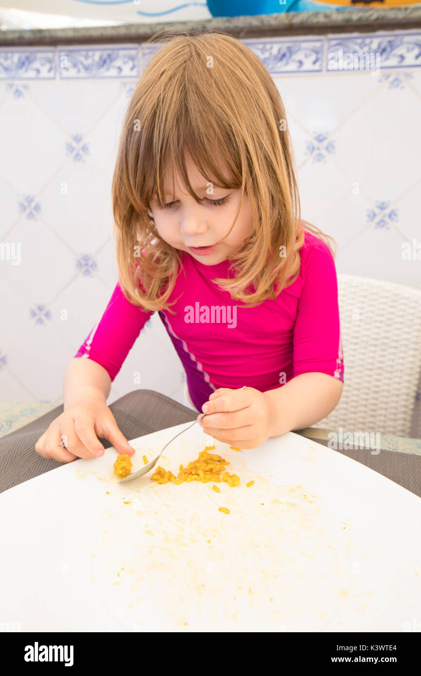 three years old blonde child with red shirt eating with spoon and finishing Spanish paella rice in big white plate Stock Photo