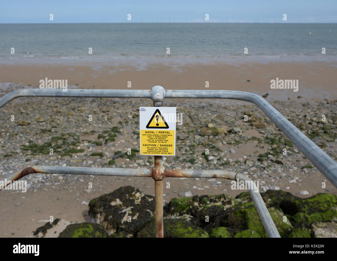 Corroded guard railing edge protection with risk warning sign and danger sign overlooking beach at low tide  in north wales uk Stock Photo