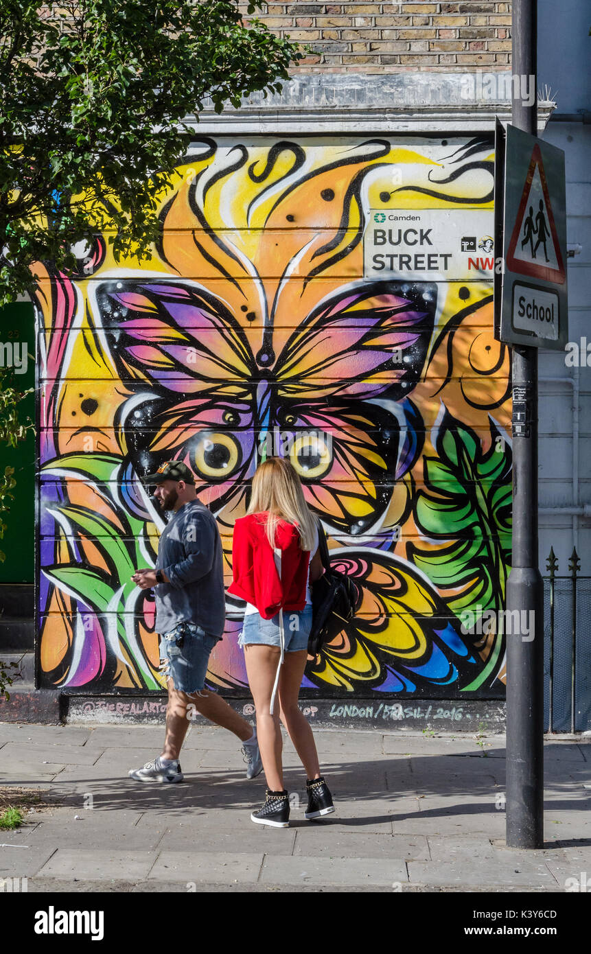 A couple of pedestrians walk past olourful street art adorns a wall on Buck Street in Camden town, London. Stock Photo