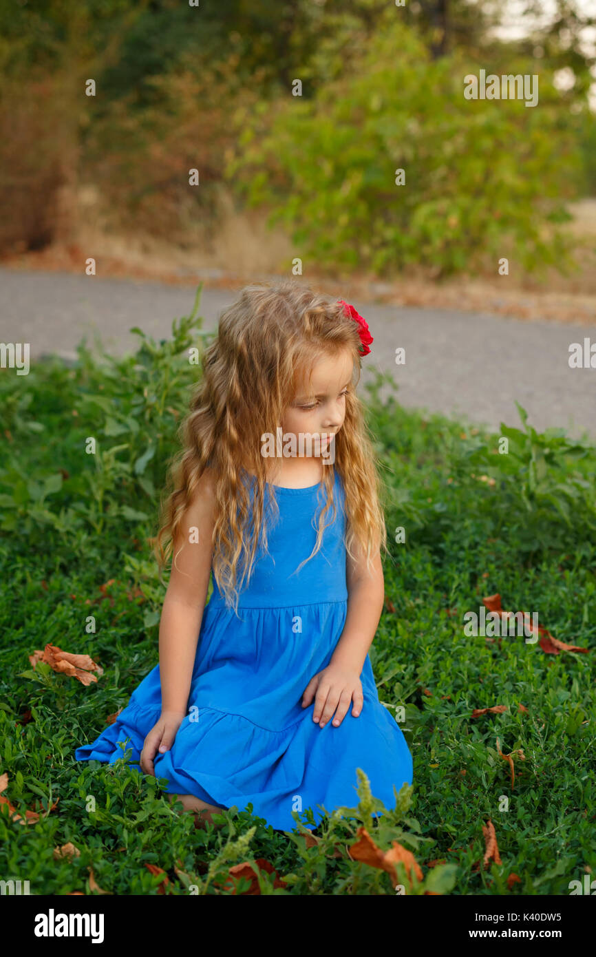 A cute little girl in a blue dress is sitting on the lawn in a city ...