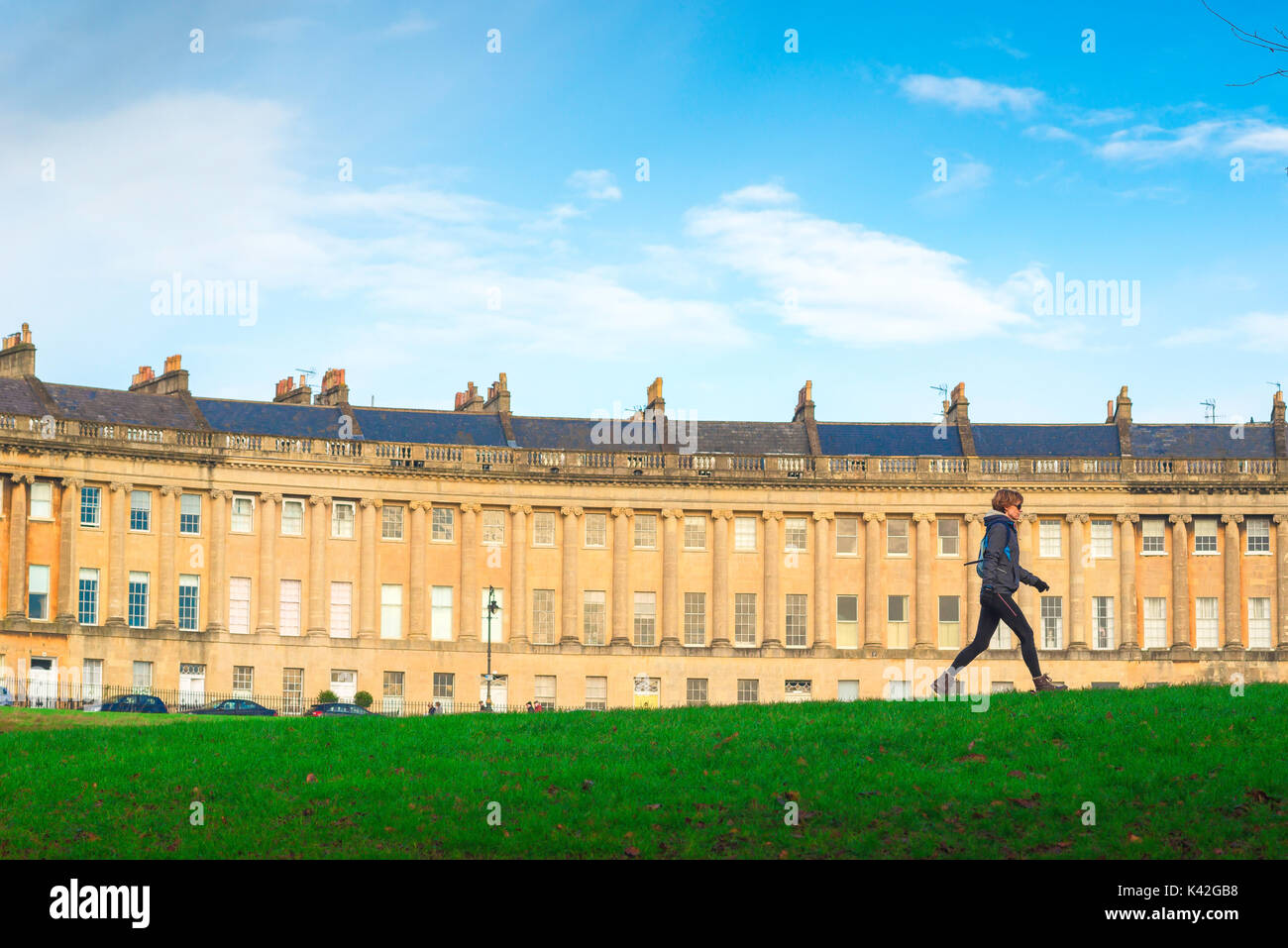 Woman walking city, against the backdrop of the Royal Crescent a solo woman walks briskly through Victoria Park in the centre of Bath, UK. Stock Photo