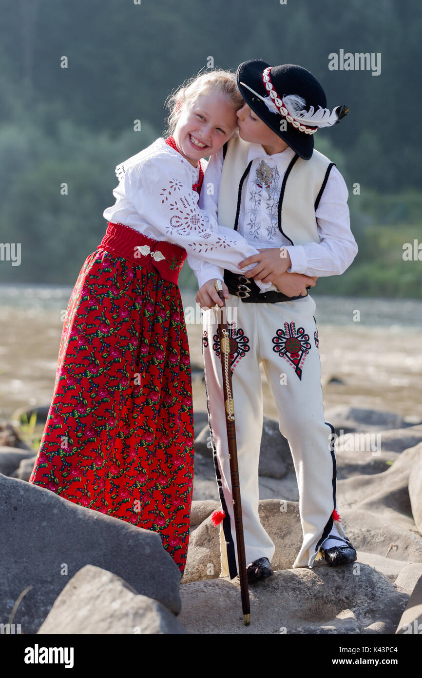 Siblings, brother spontaneously kissing smiling sister. Children wearing traditional Polish mountain people clothes standing on rocks at stream. Verti Stock Photo