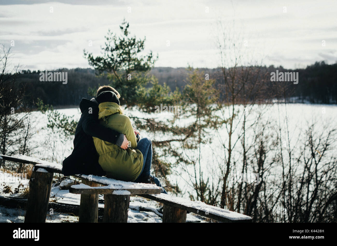 Joyful cute couple in warm clothes embraces. Young man hugs a girl. Couple sit on wooden bench in winter and look at frozen lake between forest. The c Stock Photo