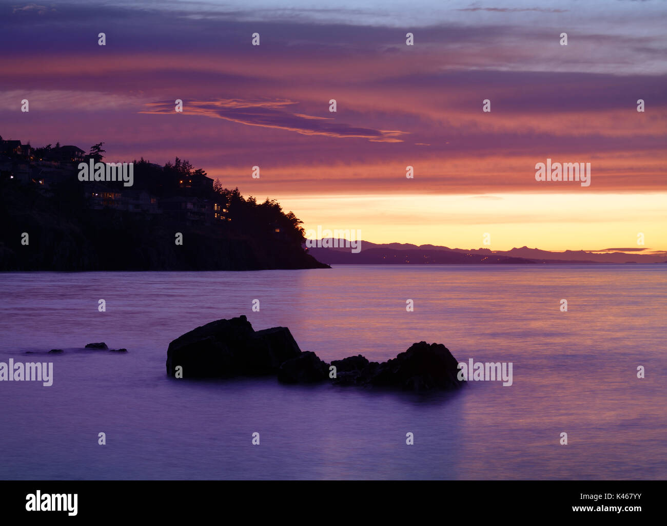 Beautiful dramatic sunset scenery in surreal pink colors on the Pacific ocean coast with lights of residential houses on dark rocky shores in Nanaimo, Stock Photo
