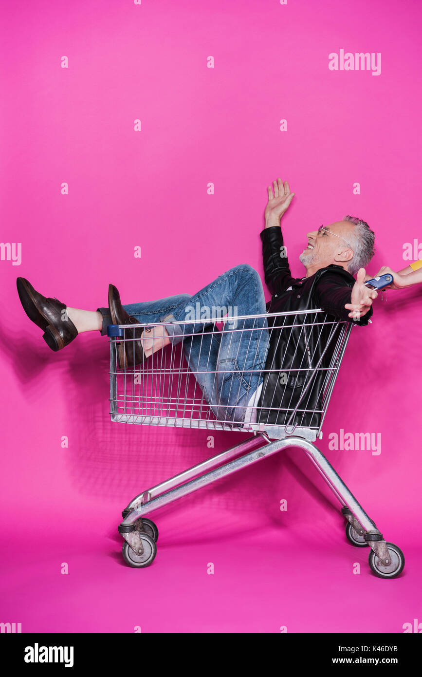 Side view of cheerful stylish senior man having fun while sitting in shopping trolley Stock Photo