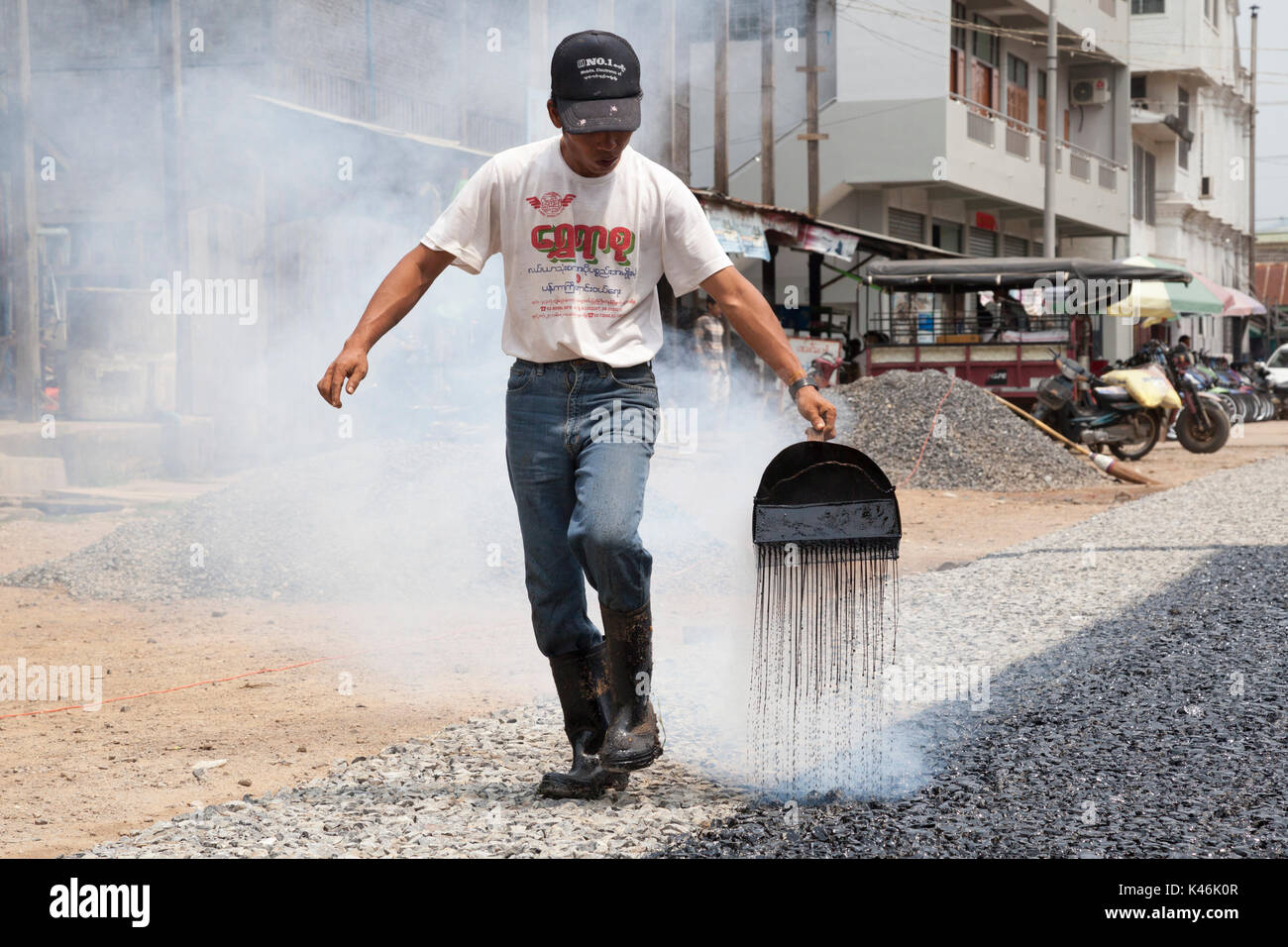 Construction worker repairing road, pouring boiling bitumen by hand onto aggregate, Hsipaw, Myanmar Stock Photo