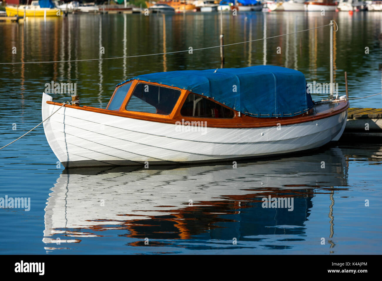Small white wooden motorboat covered with blue tarp. Boat moored at wooden pier and have rope hanging over it. Stock Photo