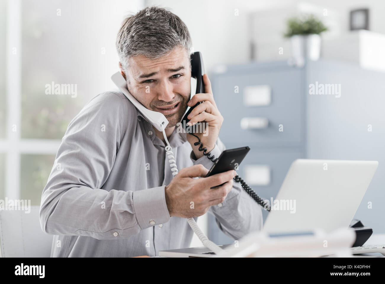 Stressed desperate businessman working in his office and having multiple calls, he is holding two handsets and a mobile phone, business management con Stock Photo