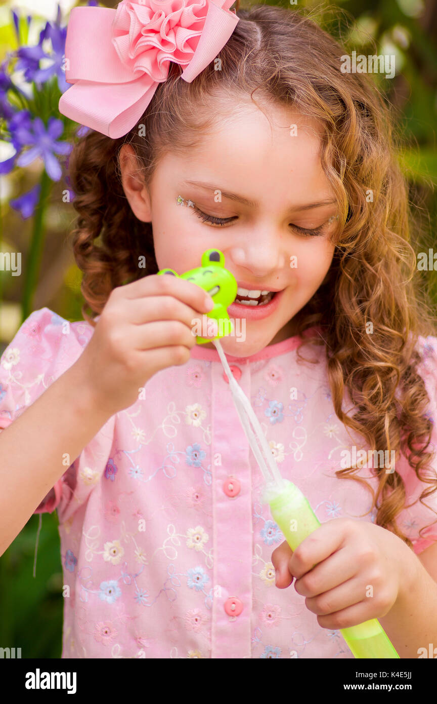 Happy little curly girl playing with soap bubbles on a summer nature, wearing a pink dress and flower in her head, in a blurred nature background Stock Photo