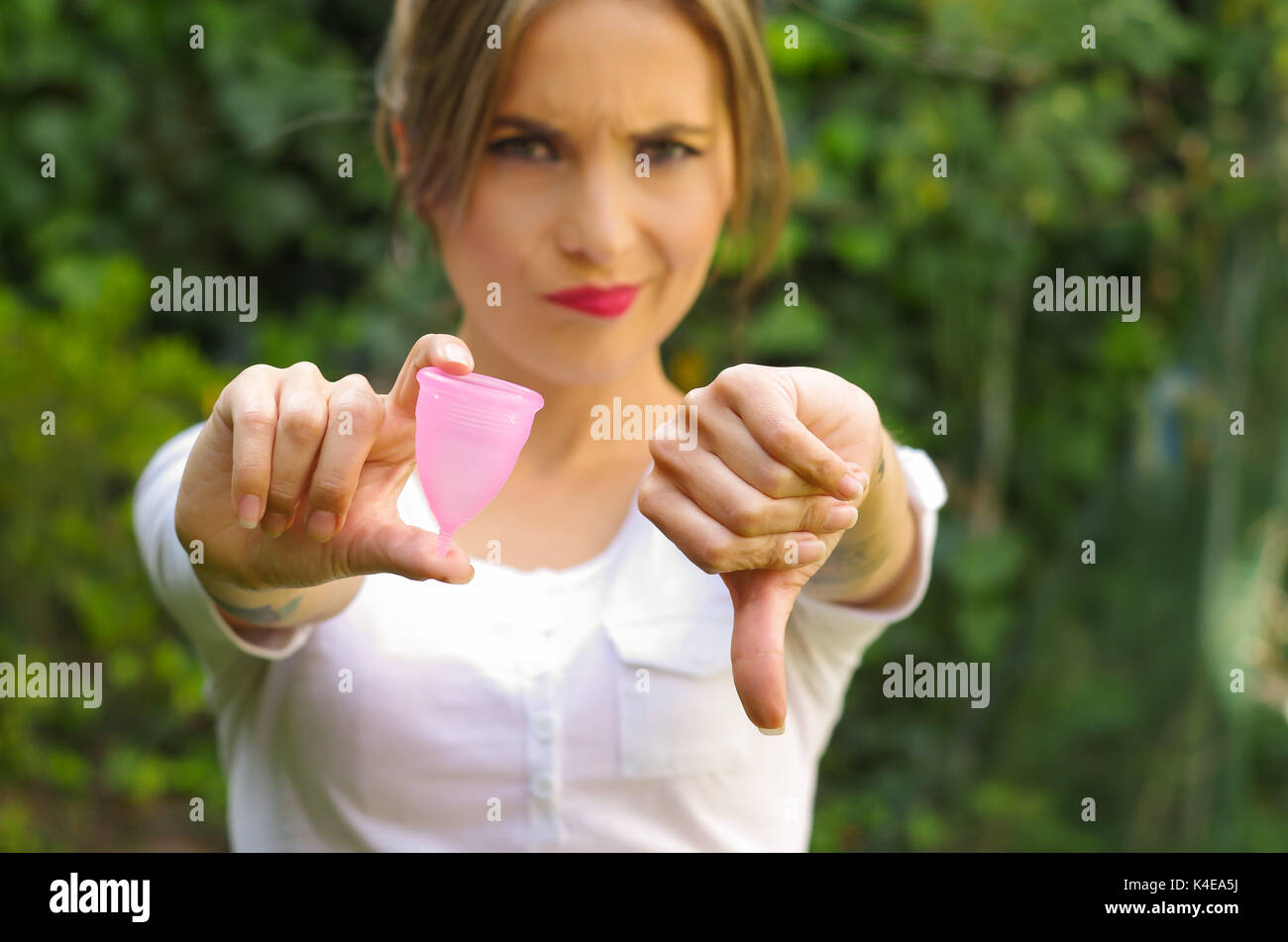 Close up of a young woman pointing in front of her a menstrual cup in one hand, Gynecology concept, ith her thums down rejecting the use of the mentrual cup, in a blurred background Stock Photo